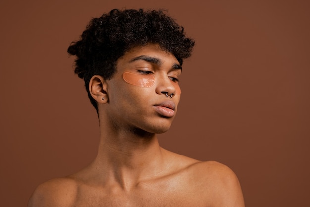 Foto de atractivo hombre negro con piercing mira hacia abajo con parches en los ojos. Torso desnudo, fondo de color marrón aislado.