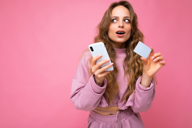 Foto de atractivo pensativo con la boca abierta joven rubia rizada vistiendo ropa rosa aislado sobre fondo de color rosa mediante teléfono móvil haciendo pagos con tarjeta de crédito mirando hacia el lado un