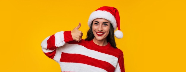 Foto de banner de gran longitud de una mujer morena linda emocionada con sombrero rojo de Navidad mientras ella está posando contra la pared amarilla