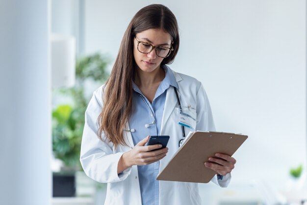 Foto de bastante doctora usando su teléfono móvil mientras revisa los informes médicos en la consulta médica.