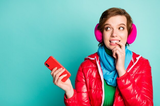 Foto de bastante hermosa dama divertida sostener el teléfono mirando interesado espacio vacío mordiendo el dedo usar abrigo rojo bufanda rosa orejeras puente verde aislado pared de color verde azulado