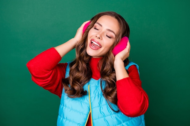 Foto de bastante linda jovencita soñadora boca abierta cerrar ojos fingir escuchar música tararear canción de cuna usar calentadores de oreja rosa chaleco azul suéter rojo aislado fondo de color verde