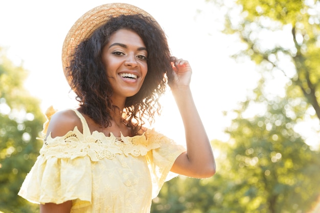 Foto de bastante mujer afroamericana de 20 años con vestido y sombrero de paja, sonriendo mientras está sentado en un banco en el parque