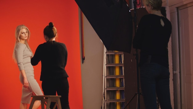 Foto entre bastidores: modelo de chica rubia juega con el pelo largo - fotógrafo toma una foto en estudio, fondo rojo