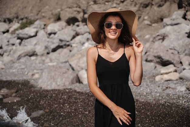 Una foto de una bella joven sensual con el pelo largo y oscuro con un vestido y un sombrero de paja posando en la playa junto al mar