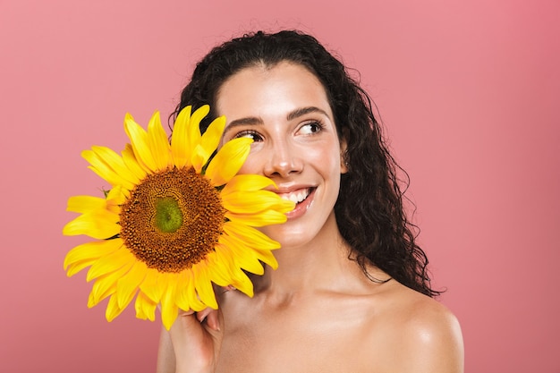 Foto de belleza de mujer desnuda caucásica de 20 años con cabello largo riendo y sosteniendo girasol amarillo, aislado sobre pared rosa