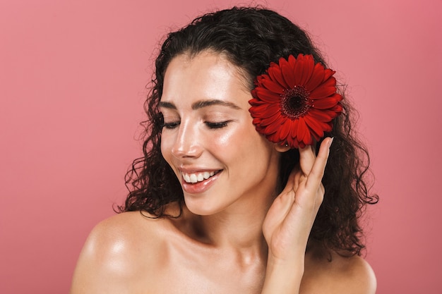 Foto de belleza de mujer desnuda europea de 20 años con cabello largo y sin maquillaje sosteniendo una flor roja, aislada sobre una pared rosa