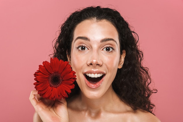 Foto de belleza de mujer semidesnuda con cabello largo y sin maquillaje sosteniendo una flor roja, aislada sobre una pared rosa