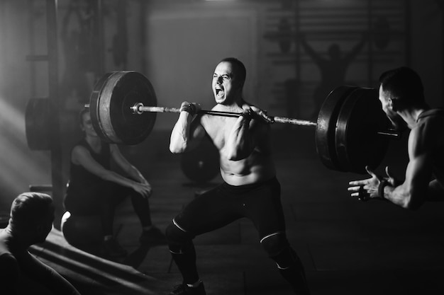 Foto en blanco y negro de deportista haciendo un esfuerzo y gritando mientras levanta pesas en entrenamiento cruzado en un gimnasio