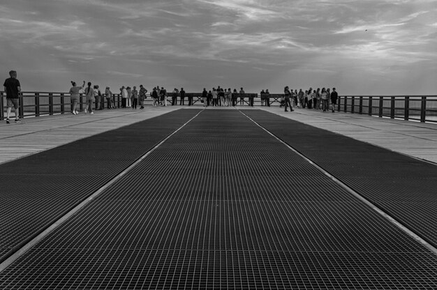 Foto una foto en blanco y negro de un muelle con gente de pie y el cielo está nublado.