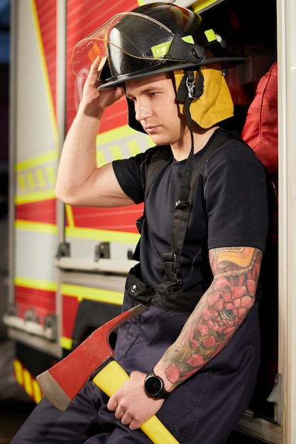 Foto de un bombero con casco con hacha contra un camión de bomberos Imagen de un joven bombero con casco en la cabeza contra el fondo de un camión de bomberos