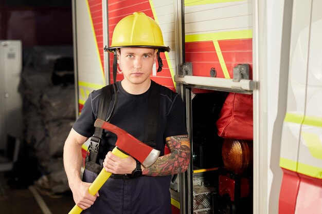 Foto de un bombero con casco con hacha contra un camión de bomberos Imagen de un joven bombero con casco en la cabeza contra el fondo de un camión de bomberos