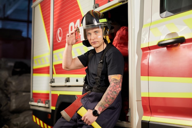 Foto de un bombero con casco con hacha contra un camión de bomberos Imagen de un joven bombero con casco en la cabeza contra el fondo de un camión de bomberos