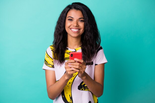 Foto de bonita dama negra mantenga smartphone brillante sonrisa mirada cámara llevar camiseta con estampado de plátano aislado de fondo de color verde azulado