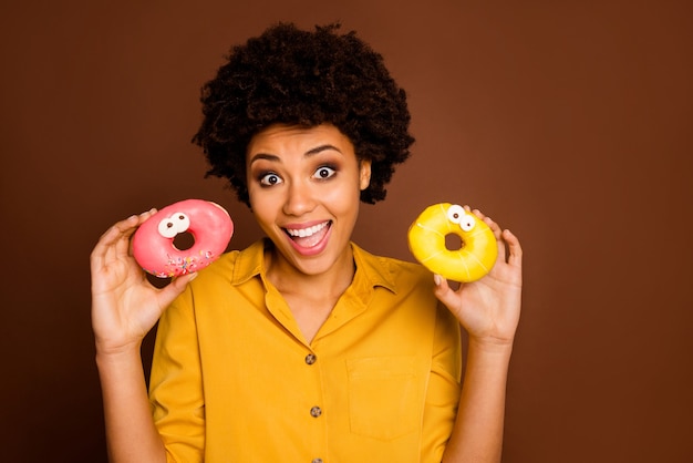 Foto de bonita dama rizada de piel oscura mantenga dos rosquillas de colores ojos caramelo rostros humanos loco despreocupado humor infantil desgaste camisa amarilla color marrón aislado