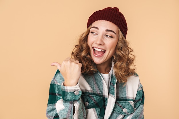 Foto de bonita mujer riendo con gorro de punto señalando con el dedo y mirando a un lado aislado en beige