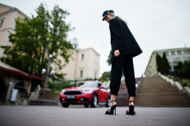 Foto borroneada de modelo de mujer de moda sexy rubia hermosa en gorra y en todo negro con maquillaje brillante cerca del coche rojo de la ciudad.