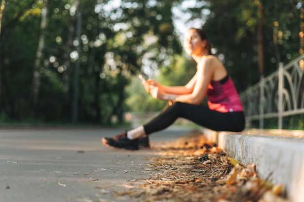 Foto borrosa de una joven morena delgada con ropa deportiva descansando y usando un teléfono móvil en el parque de otoño en la hora dorada del amanecer Salud y bienestar estilo de vida