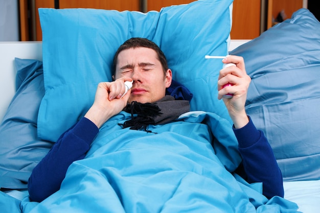 Foto de brunet enfermo en bufanda con pañuelo y spray con medicina acostado en la cama