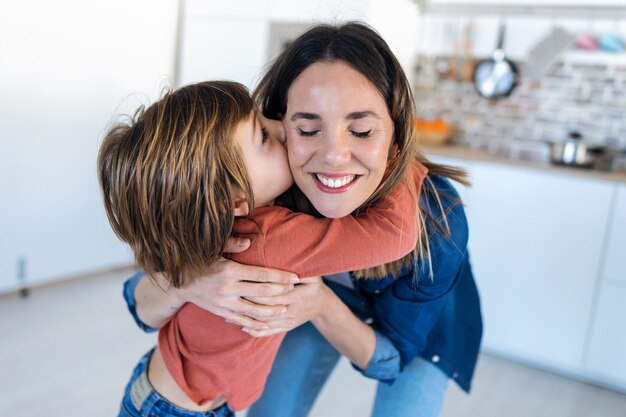 Foto de buen chico besando a su madre mientras está de pie en la cocina de su casa.