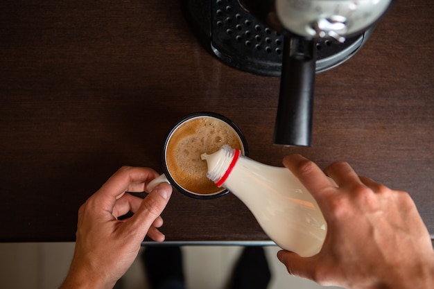 Foto de cafetera, manos del hombre vertiendo leche en una taza de café en la cocina