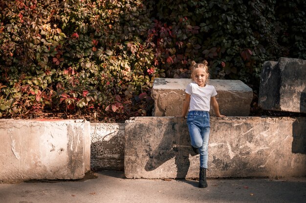 Foto de la calle de una hermosa niña en jeans y una camiseta blanca sobre un fondo de losas de hormigón y hojas de otoño