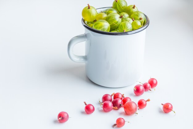 Foto cambiante de grosellas verdes jugosas en una taza blanca de pie sobre un fondo gris con algunas bayas por ahí tomadas con luz natural