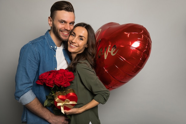 Foto foto de cerca de un hombre atractivo, que sostiene una caja roja en sus manos, mientras que su hermosa novia le sonríe después de recibir un ramo de rosas.