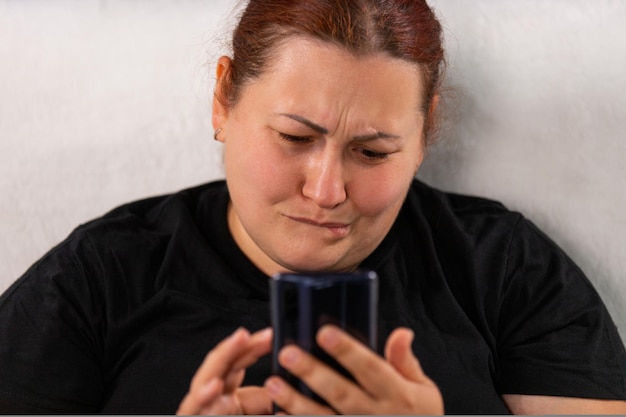 Foto de cerca de una mujer con expresión facial triste en camiseta negra sosteniendo el teléfono móvil en su ...