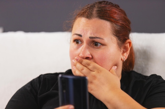 Foto de cerca de una mujer preocupada en camiseta negra sentada en el sofá blanco sosteniendo el teléfono inteligente ...