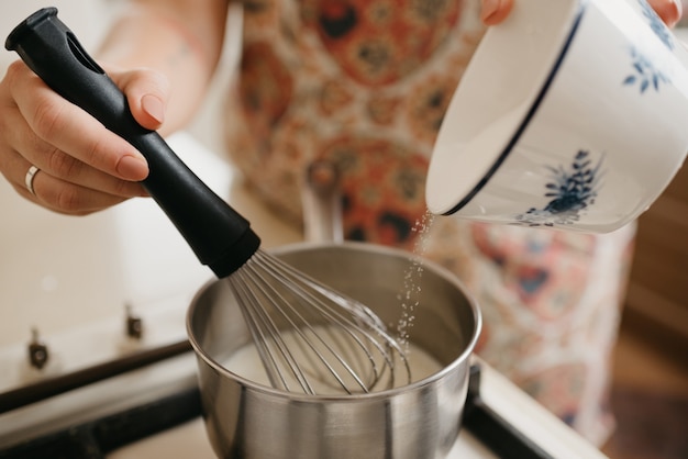 Una foto cercana de una mano de una mujer joven mezclando la crema de corola, la ralladura triturada, el jugo de limón y agregando azúcar en la cacerola de la estufa de gas