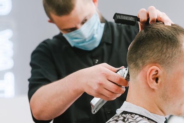 Foto cercana del proceso de corte de pelo de cuarentena, el peluquero usa una máscara médica en la cara, un peluquero profesional con una máscara protectora corta el cabello del cliente con una cortadora