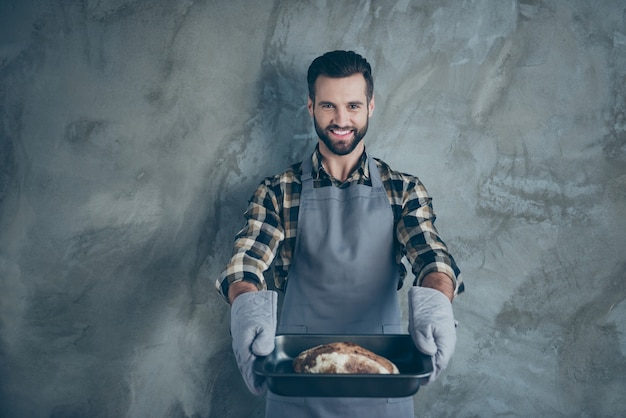 Foto de chef cocinero positivo alegre habiendo cocinado su pan de marca sirviéndole esto en guantes pared aislada pared de color gris de hormigón