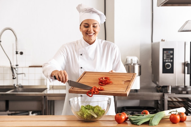 Foto de chef mujer europea vistiendo uniforme blanco haciendo ensalada con verduras frescas, en la cocina del restaurante