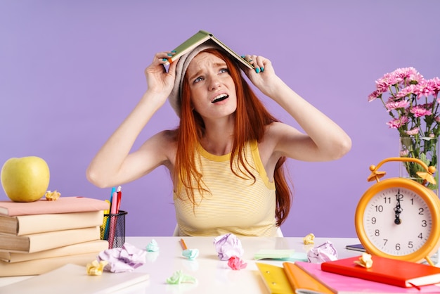 Foto foto de chica estudiante confundida sosteniendo el libro de ejercicios sobre su cabeza mientras hace la tarea aislada sobre la pared púrpura