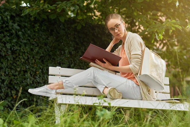 Foto de una chica joven, bonita y de moda, que está leyendo un libro en el banco del parque blanco. Viste ropa de colores claros: jeans, camiseta y un cárdigan. También lleva mochila y anteojos.