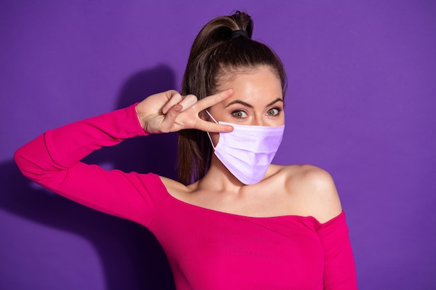 Foto de chica loca positiva hacer v-sign cerca de la cara usar máscara médica aislada sobre fondo de colores vivos