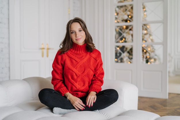 La foto de una chica tranquila y despreocupada usa poses cálidas de ropa doméstica en un sofá blanco mira directamente a la cámara pasa el fin de semana de invierno en casa anticipa el concepto de vacaciones de abeto decorado de Año Nuevo