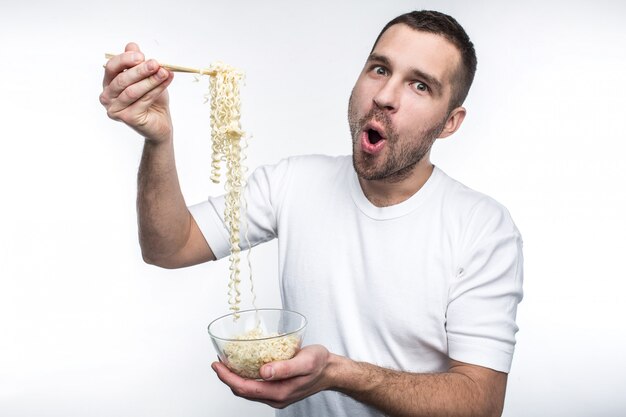 Una foto de un chico al que le gusta comer cocina oriental. Tiene un plato de espagueti tailandés y lo guarda con palitos especiales para comer. También a este tipo raro le gusta comer comida chatarra. Aislado en la pared blanca