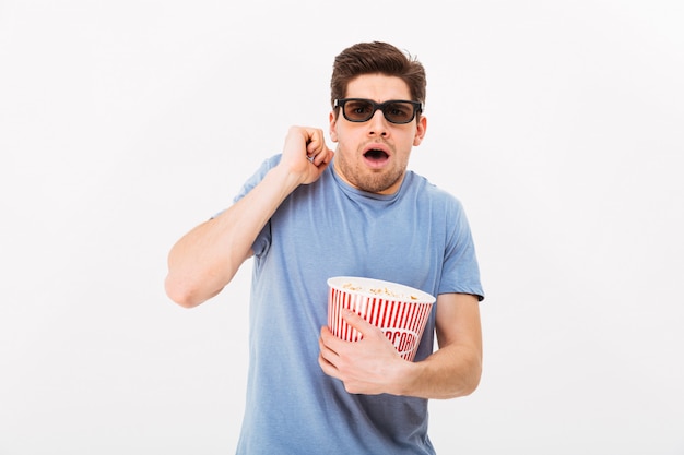 Foto de chico caucásico en camiseta casual y gafas 3d comiendo palomitas de maíz y viendo películas emocionalmente en el cine, aislado sobre la pared blanca