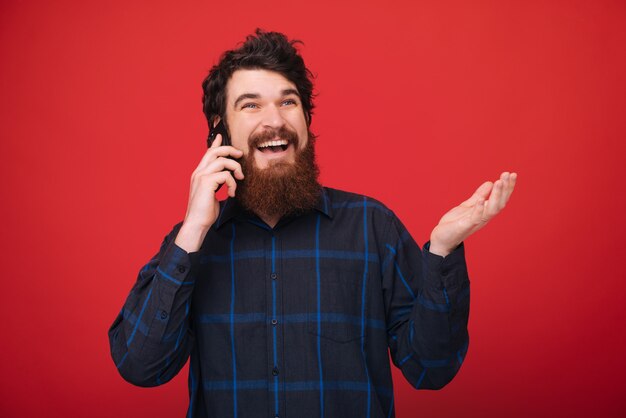 Foto de chico guapo con barba hablando por teléfono sobre pared roja