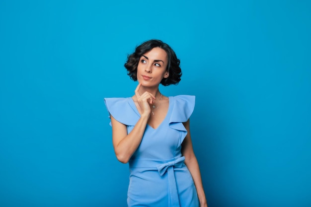 Foto de cierre de una encantadora mujer sana con un vestido azul que mira a la cámara y sonríe mientras está de pie y posa aislada en el fondo azul