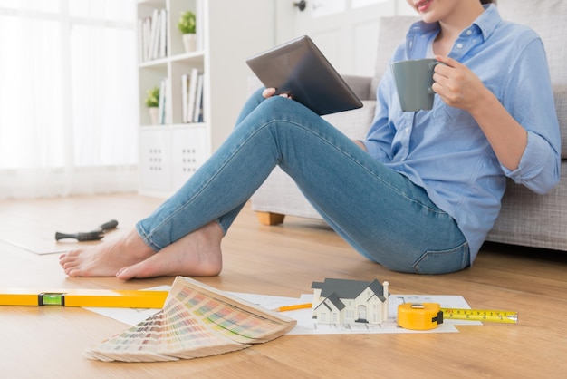 foto de cierre de una mujer hermosa bebiendo café caliente usando una computadora portátil y haciendo trabajos de diseño interior de la casa con una herramienta de ingeniería en el piso de madera de la sala de estar.