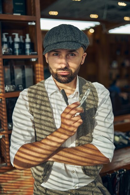 Foto de cintura para arriba de un barbero decidido con un sombrero sosteniendo una navaja de cortar el cuello mientras mira a la cámara