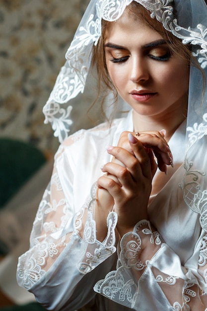 Foto close-up de uma noiva com maquiagem profissional, ela está com um jaleco branco, véu no rosto. Dia do casamento da jovem. Sessão de fotos, penteado, beleza