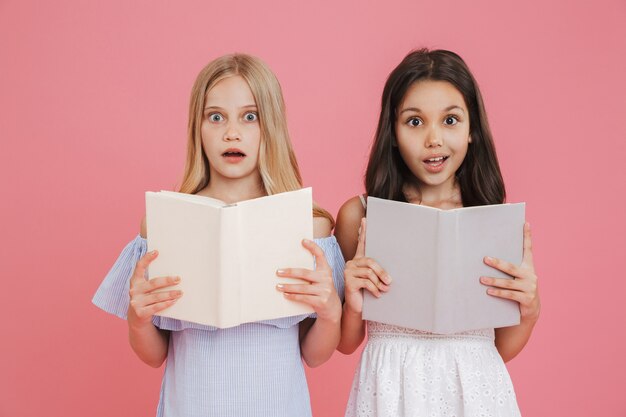 Foto de colegiala morena y rubia con vestidos sosteniendo y leyendo libros junto con entusiasmo, aislado sobre fondo rosa