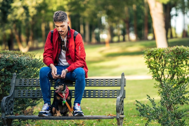 Foto com cópia de um cachorro e um homem sentado em um banco de um parque usando um celular