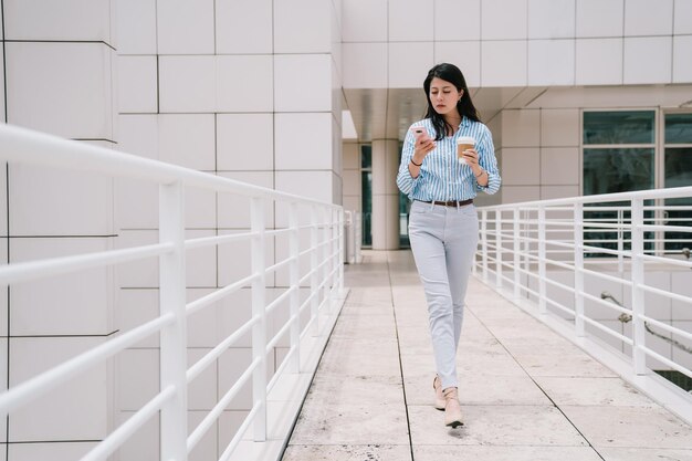 una foto completa de una dama elegante que usa el teléfono mientras camina por el puente, sostiene una taza de café