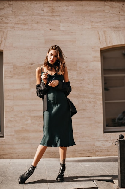 Foto completa de jovem bem construída com cabelos escuros e lábios vermelhos em vestido midi verde jaqueta preta e sapatos em pé e posando contra o fundo do edifício claro