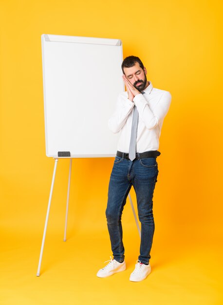 Foto completa do empresário dando uma apresentação no quadro branco sobre fundo amarelo isolado, fazendo o gesto do sono em expressão dorable